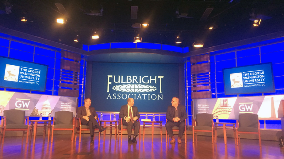 Three men sit on stage with Fulbright Association graphic on screen in background