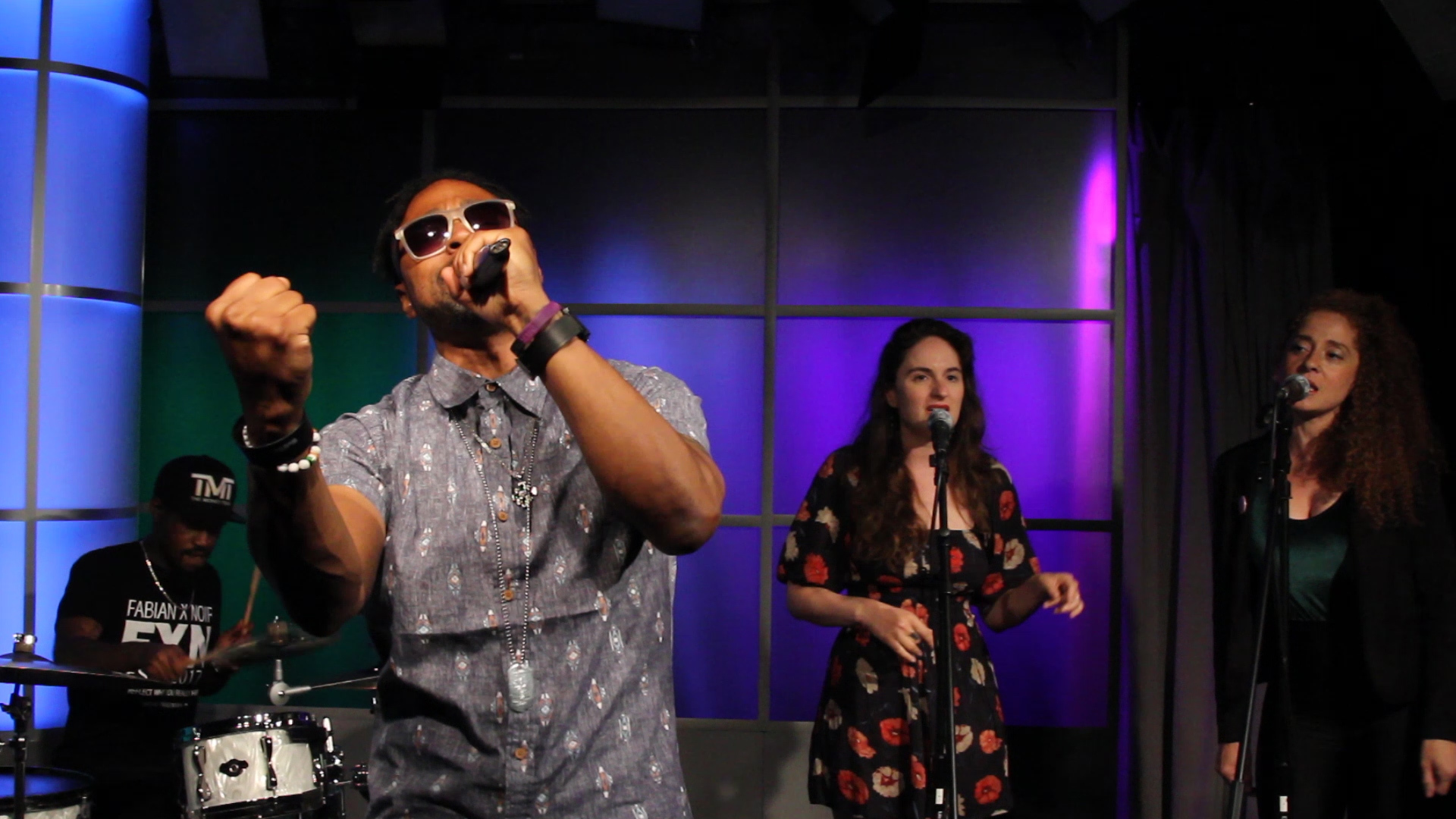 Young man holding a mic wearing sunglasses and a band behind him on a stage