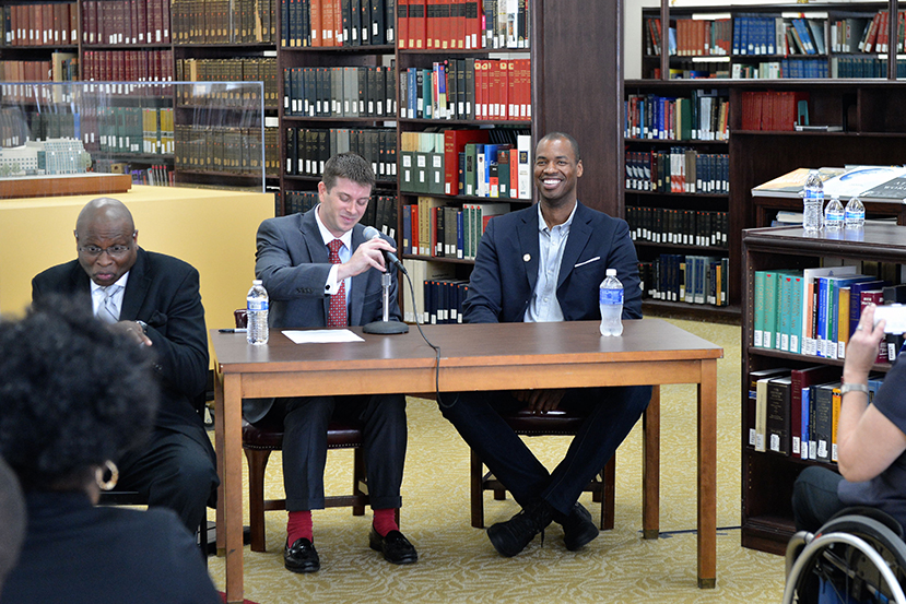 Three men sit at a table in a library in front of a small audience