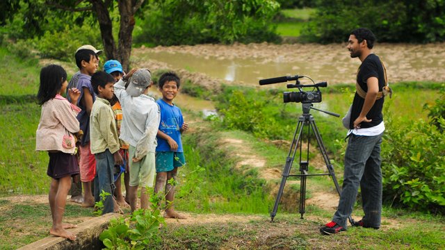 Man stands in front of group of young Cambodian children with video camera on tripod. 