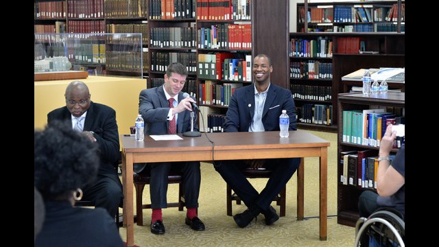 Three men sit at a table in a library in front of a small audience