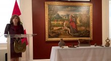 Woman stands at podium with large painting on a wall beside her and artifacts on a table