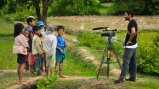 Man stands in front of group of young Cambodian children with video camera on tripod. 