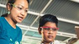 Woman wearing construction goggles stands beside young boy also wearing goggles, both looking at an object