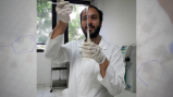 Man in lab coat holds sticks above a vile that has long clear sticky substance hanging from the stick into the vile 