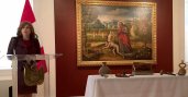 Woman stands at podium with large painting on a wall beside her and artifacts on a table